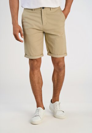 Beige cotton shorts with a rolled hem. Front pockets and a single button closure. Worn with white sneakers, standing on a neutral background.