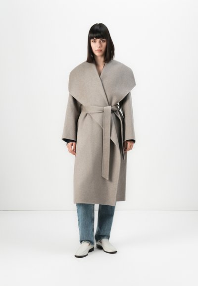 Femme avec des cheveux noirs raides portant un manteau en laine beige ceinturé sur un jean bleu et des chaussures blanches, se tenant devant un fond blanc.