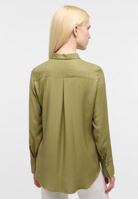 Camicia verde oliva a maniche lunghe realizzata in tessuto morbido, dotata di yoke posteriore e polsini con bottone, con un orlo curvo.
