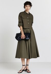 Donna con camicia verde oliva bottonata e gonna midi ampia, che tiene una borsa a mano nera e indossa sandali neri con tacco a strappo, su sfondo bianco.