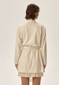 Kremowy szlafrok z teksturowanym wzorem diamentów, długimi rękawami oraz paskiem w talii, sięgający do długości do połowy uda.