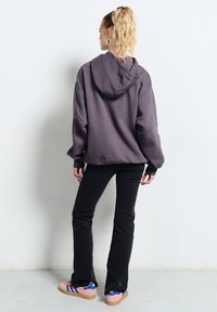 Kobieta z kręconymi włosami związanymi w kucyk, ubrana w fioletową bluzę z kapturem, czarne spodnie i różowe trampki, stoi tyłem do białej ściany.