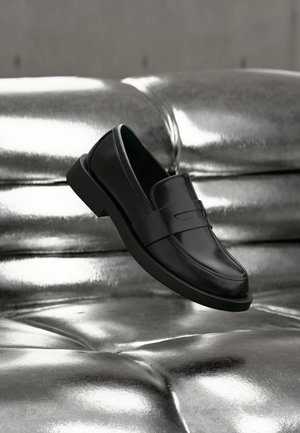 Pantof din piele neagră, tip loafer, plutind deasupra unei suprafețe lucioase, tușate, argintii metalice.