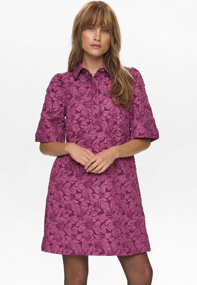 Rochie fucsia cu un model floral texturat, mâneci scurte, guler și o siluetă evazată, asortată cu colanți întunecați.