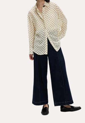 Personne portant un chemisier crème à manches longues à pois, un jean large foncé et des mocassins noirs, debout avec une main dans le dos.