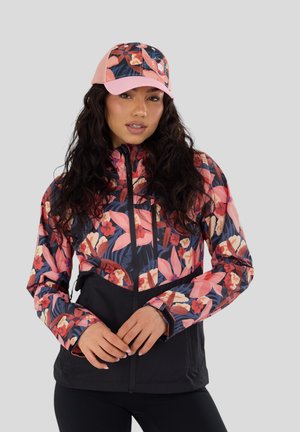 Donna con capelli lunghi e scuri che indossa una giacca nera con motivo floreale rosa e un berretto rosa floreale abbinato, in piedi davanti a uno sfondo grigio.