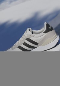 Baskets Adidas blanches et noires avec trois bandes, design lacé et semelle coussinée sur un fond bleu et blanc flou.
