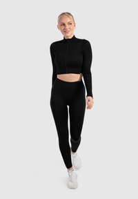 Sort ribbet crop top med høj krave og lynlås foran, parret med højtaljede sorte leggings. Modellen bærer hvide sneakers.