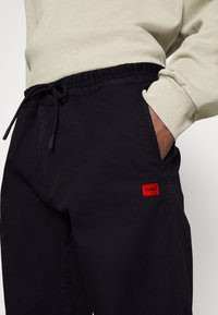 Person som bär svarta dragkedjeshorts med en röd HUGO-logotyp på fickan och en beige långärmad tröja, med handen i fickan.