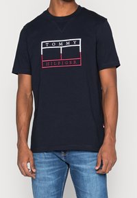 Muž v námořnickém modrém tričku s krátkým rukávem s bílým a červeným logem Tommy Hilfiger, v kombinaci se středně modrými džínami.