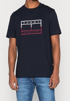 Hombre vestido con una camiseta de manga corta azul marino con el logo de Tommy Hilfiger en blanco y rojo, combinada con unos vaqueros de lavado medio.