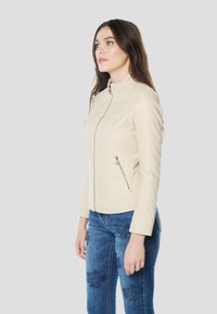 Beige neplederen motorjas met opstaande kraag, asymmetrische ritssluiting aan de voorkant en doorgestikte details op de schouders en mouwen.