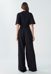 Ženska z dolgimi rjavimi lasmi stoji obrnjena proč, oblečena v črn enodelni kombinezon s širokimi hlačnicami in kratkimi rokavi, proti enobarvni svetli ozadju.