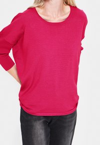 Kvinna i ljusrosa långärmad tröja med ribbade ärmslut och urtvättrade svarta jeans, med händerna vilande bakom ryggen.