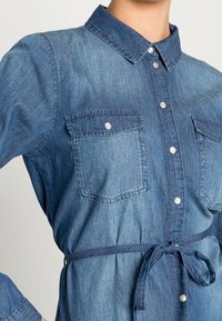 Denimskjorta i medelblått med krage, knäppning framtill, två bröstfickor och bältad midja. Mjuk textur med en avslappnad design.