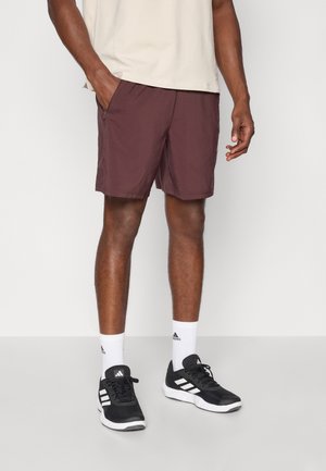 Homme portant un short marron foncé, une chemise beige, des chaussettes blanches Adidas et des baskets noires Adidas, debout avec une main dans la poche.