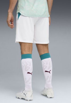 Męskie nogi w białych szortach piłkarskich, białych korkach i białych podkolanówkach z turkusowymi ściągaczami oraz ciemnymi logo Puma, stojące na szarej podłodze.