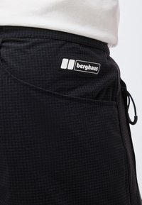 Κοντινό πλάνο μαύρου καρό παντελονιού με πλαϊνή τσέπη και λογότυπο Berghaus, φορεμένο με λευκό τοπ.