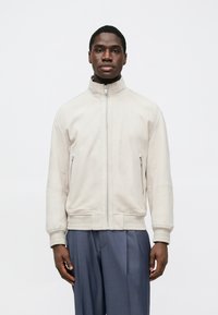 Veste zippée beige clair en matière douce avec poignets et ourlet côtelés, dotée de deux poches latérales et d'un col montant.
