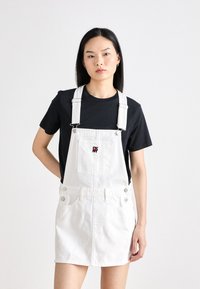 Belo jeans overall obleka z nastavljivimi naramnicami, sprednjim žepom in okrasnimi gumbi ob straneh. Nosijo jo čez črno majico.