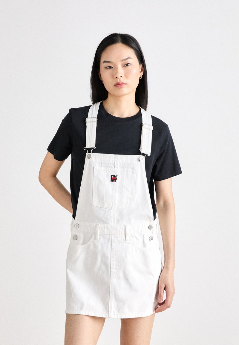 Belo jeans overall obleka z nastavljivimi naramnicami, sprednjim žepom in okrasnimi gumbi ob straneh. Nosijo jo čez črno majico.