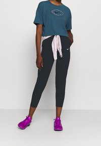 Turkis cropped t-shirt med en blomstret Nike-logo, kombineret med sorte leggings. Lyserød top bundet om taljen og lyse lilla træningssko.