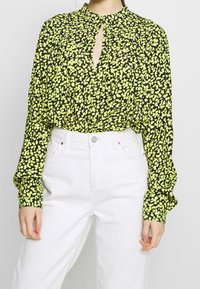 Černá blůza s žlutým květinovým vzorem, dlouhými rukávy, detaily ve tvaru klíčové dírky na výstřihu a staženými manžetami, nošena s bílými kalhotami s vysokým pasem.