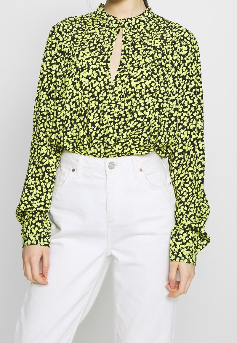 Černá blůza s žlutým květinovým vzorem, dlouhými rukávy, detaily ve tvaru klíčové dírky na výstřihu a staženými manžetami, nošena s bílými kalhotami s vysokým pasem.