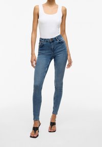 Hoge taille blauwe spijkerbroeken met een getailleerde silhouet, vijf zakken en een knoopsluiting, gecombineerd met een witte tanktop en zwarte sandalen.