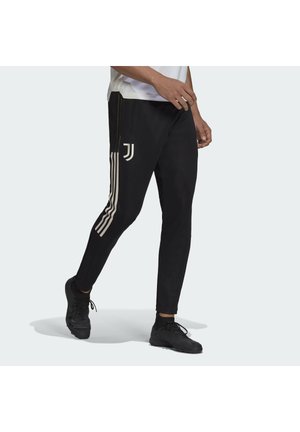 Homme marchant portant un pantalon de sport noir avec des bandes blanches et un logo de la Juventus sur la cuisse, associé à des baskets noires.