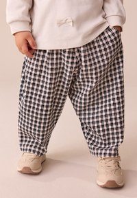 Chemise blanche à manches longues avec un petit détail en nœud, associée à un pantalon large à carreaux bleu marine et blanc et des baskets beiges.