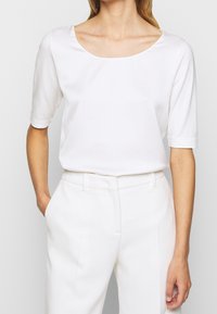 Blouse blanche à manches courtes avec un large col rond et une texture lisse, associée à un pantalon blanc ajusté, doté de poches latérales.