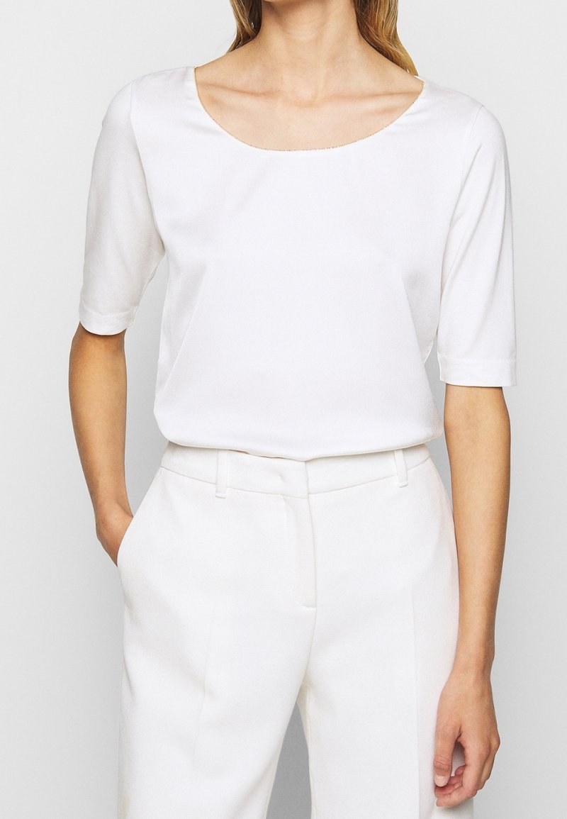 Blouse blanche à manches courtes avec un large col rond et une texture lisse, associée à un pantalon blanc ajusté, doté de poches latérales.