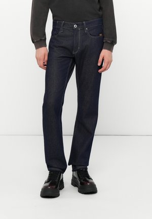 Homme portant un jean droit bleu foncé, des chaussures noires épaisses à lacets et un haut à manches longues foncé sur fond blanc.