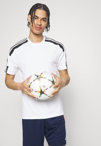 Vitt idrottströja med svarta ränder, som matchas med mörkblå shorts. Personen håller en flerfärgad fotboll med stjärnmönster.