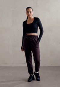 Schwarzes Langarm-Crop-Top kombiniert mit dunkelbraunen Jogginghosen. Schwarze Sneaker mit weißen Akzenten runden das Outfit ab. Glatte Texturen überall.