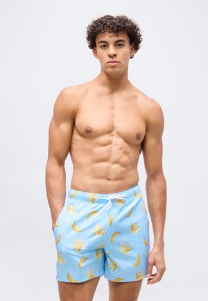 Młody mężczyzna o kręconych włosach, ubrany w jasnoniebieskie kąpielówki z żółtymi nadrukami bananów, stojący na tle jednolitego białego tła.