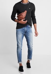 Suéter negro de punto con cuello de marca, jeans azul claro ajustados y zapatillas negras. Bolso de cuero marrón tipo bandolera con detalles de cremallera.