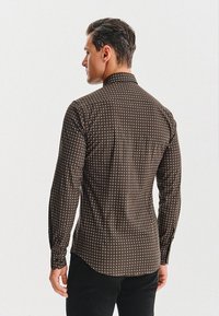 Mężczyzna zwrócony plecami, ubrany w dopasowaną koszulę z długim rękawem o małym geometrycznym wzorze w kolorze brązowym i czarnym oraz czarne spodnie.