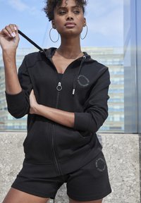 Haut à zip noir et shorts assortis en tissu texturé. Le hoodie présente un cordon de serrage et un détail de logo, avec une coupe décontractée.