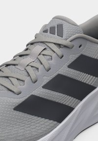 Κοντινό πλάνο γκρι αθλητικού παπουτσιού Adidas με μαύρες ρίγες, ύφασμα από δίχτυ, δεμένα κορδόνια και μαλακή λευκή σόλα.