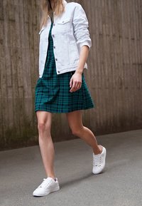 Femme marchant vêtue d'une veste blanche, d'une robe à carreaux verts et de baskets blanches sur un chemin en béton près d'un mur texturé.