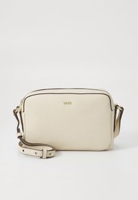 Crèmekleurige leren crossbodytas met een gladde textuur, bruine bies, goudkleurig logo en een verstelbare riem. Rechthoekige vorm met ritssluiting.