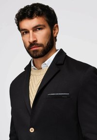 Schwarzer Blazer mit Schößchen, einem einzelnen Holzknopf und einer Brusttasche mit einem gemusterten Akzent. Beiger Strickpullover darunter.