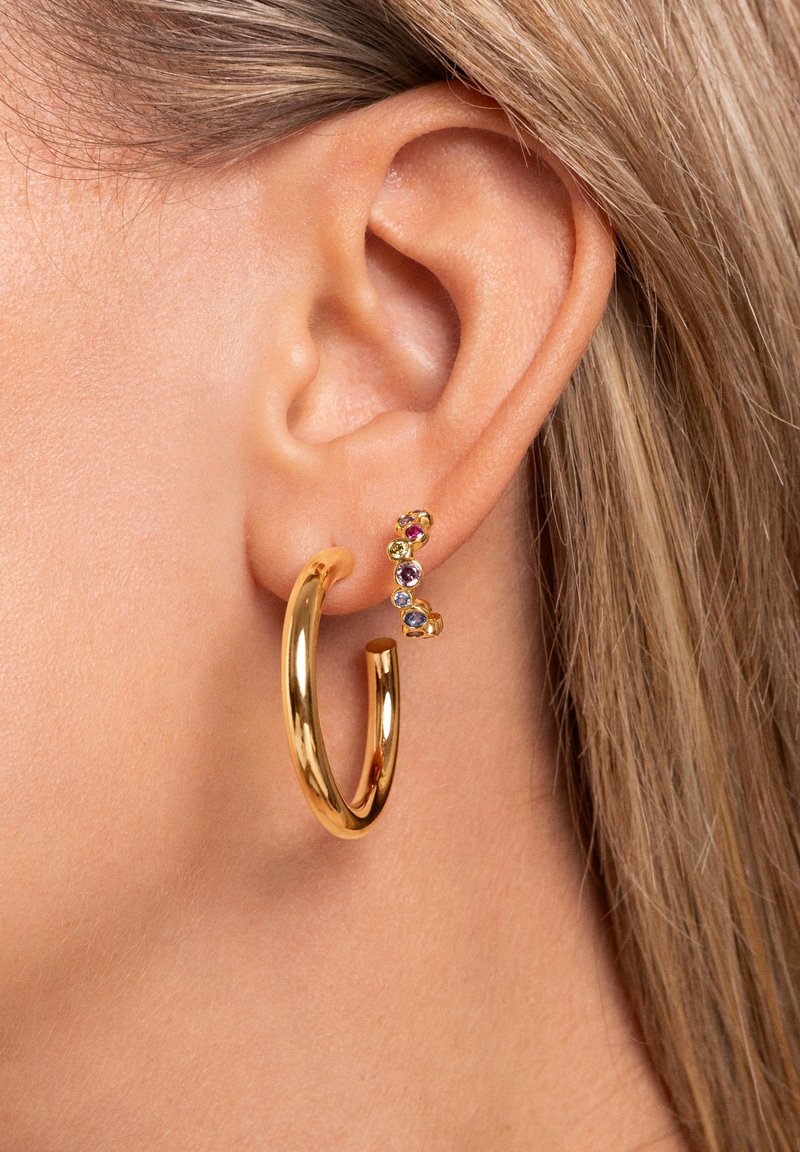 Gros plan d'une oreille avec deux boucles d'oreilles en or : un grand anneau simple et une petite barre courbée ornée de pierres précieuses multicolores.