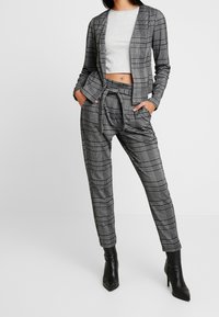 Šedý a černý kostkovaný oblek na míru s crop topem, který se skládá z dobře padnoucího blejzru a kalhot s vysokým pasem. Černé kotníkové boty na podpatku.