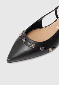 Zapato plano negro con punta puntiaguda, tira trasera y tachuelas doradas decorativas a lo largo del borde superior, con suela interior beige.