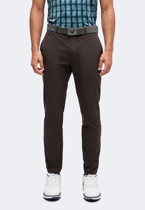 Homme portant un pantalon slim noir, une ceinture grise avec une boucle foncée, des baskets blanches et une chemise bleue à carreaux rentrée.