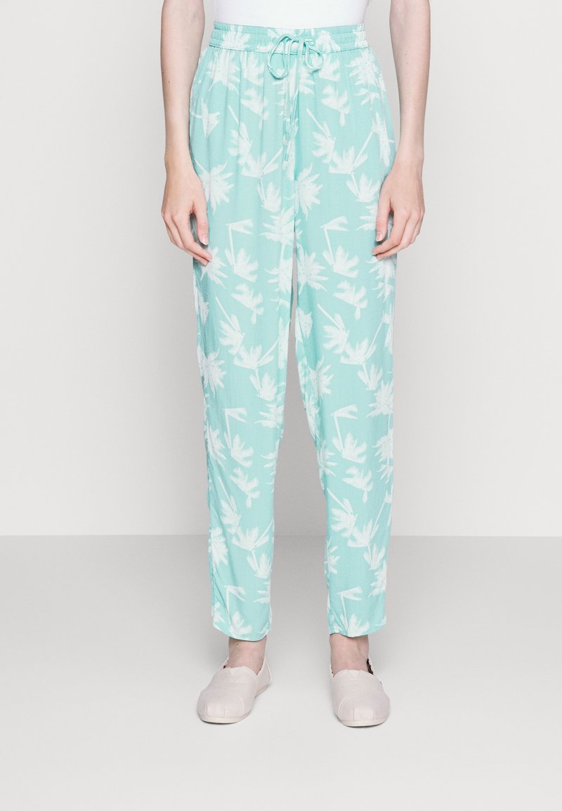 Person klädd i ljusblå byxor med vitt blommönster och dragsko i midjan, kombinerat med ljusa rosa slip-on skor.