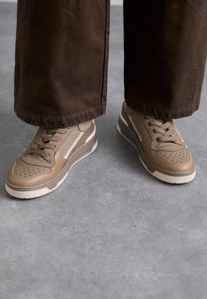 Persoon die bruine broek met wijde pijpen en beige vetersneakers draagt, staand op een grijze vloer.